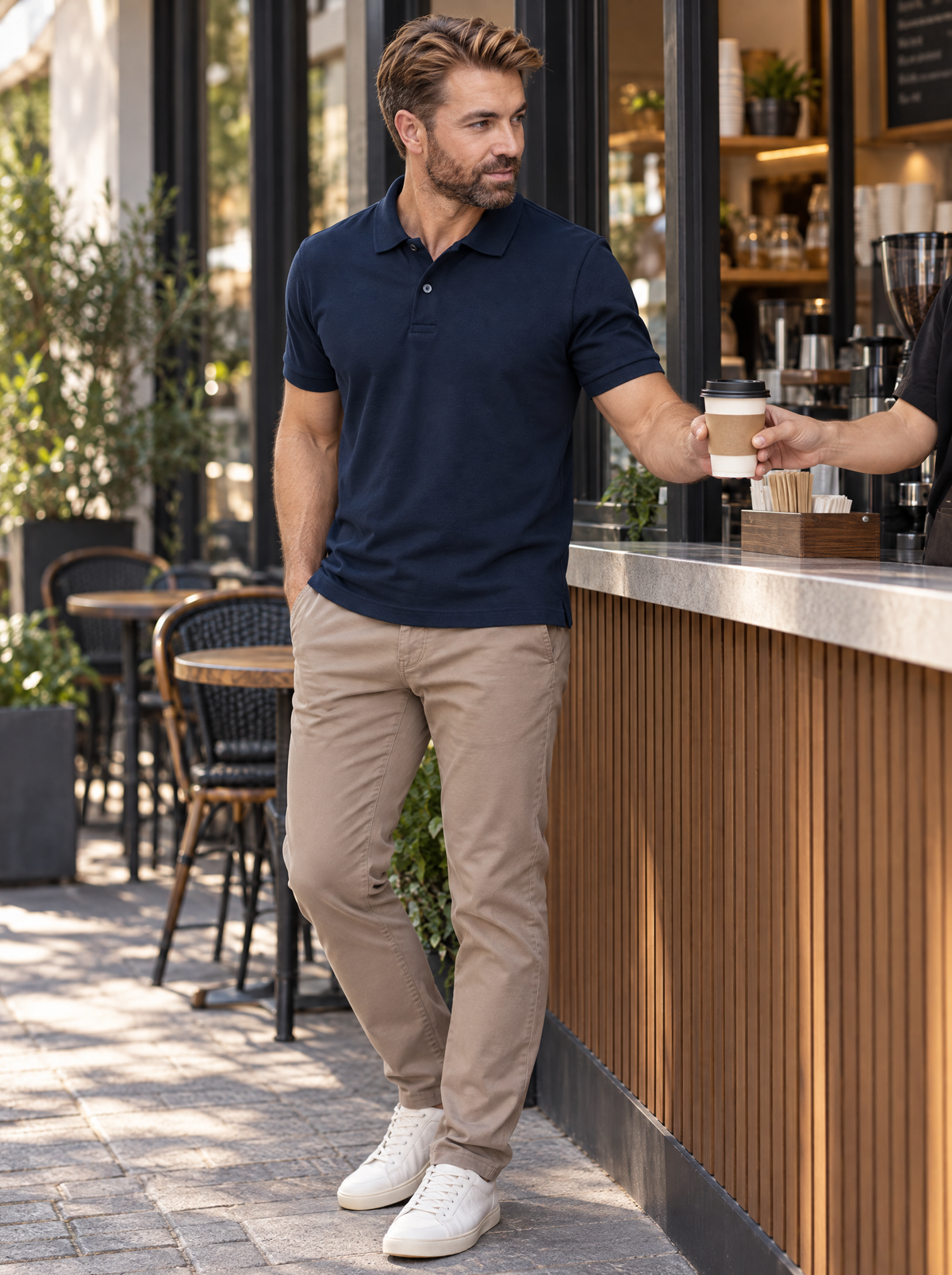 Homem em pé no balcão de um café, vestindo polo marinho e calça chino caqui